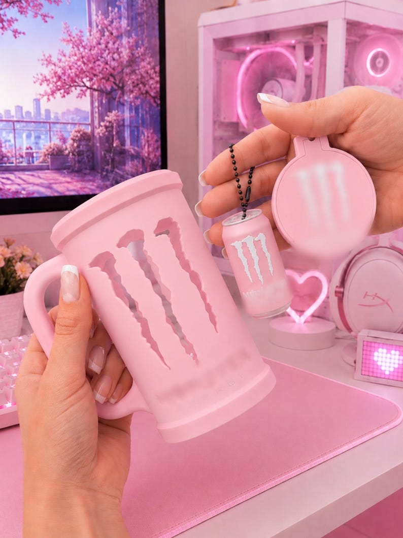 May include: A light pink mug featuring a large cutout of the Monster Energy logo on the side. Accompanying the mug is a small, matching light pink miniature can keychain and a circular light pink compact mirror, both displaying the Monster logo.