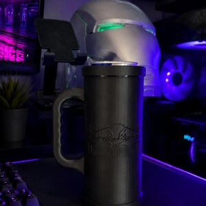 May include: A matte black cylindrical beverage can holder featuring a sturdy side handle and a debossed Red Bull logo on the front. The holder is designed to securely fit a standard beverage can, which is visible at the top of the container.