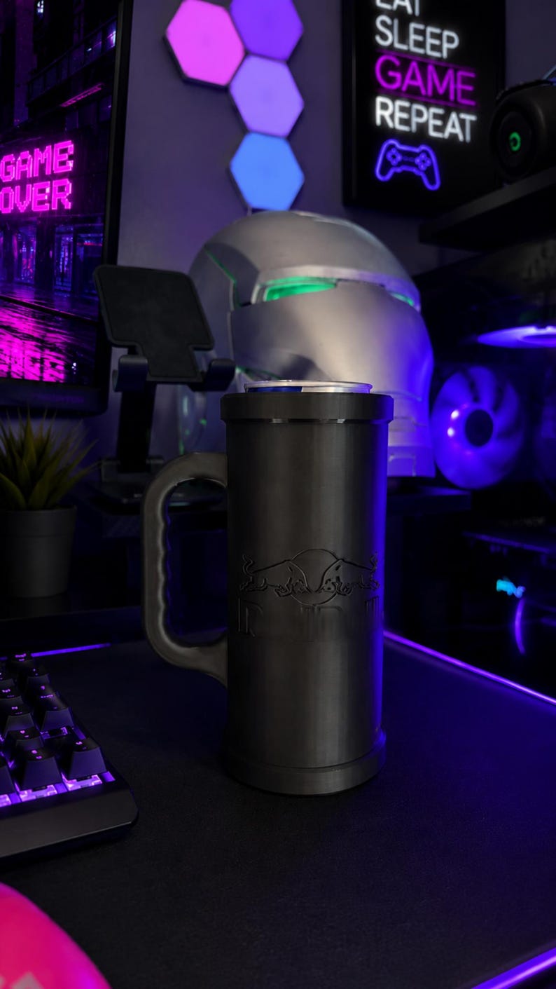 May include: A matte black cylindrical beverage can holder featuring a sturdy side handle and a debossed Red Bull logo on the front. The holder is designed to securely fit a standard beverage can, which is visible at the top of the container.