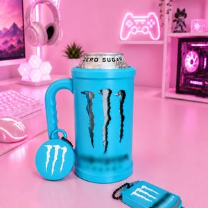 May include: A bright blue cylindrical beverage holder with a handle, featuring a black claw mark graphic and the Monster logo. A silver can labeled Zero Sugar is inserted inside. A circular tag and a rectangular tag with the logo are attached.