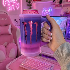 May include: A cylindrical beverage can holder with a handle, featuring a gradient purple and pink exterior. The holder has a cutout design revealing a pink Monster Energy drink can inside, with the iconic claw logo visible through the opening.