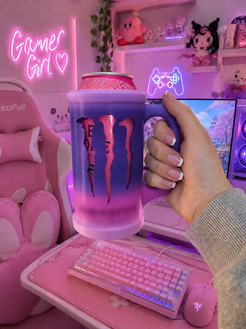May include: A cylindrical beverage can holder with a handle, featuring a gradient purple and pink exterior. The holder has a cutout design revealing a pink Monster Energy drink can inside, with the iconic claw logo visible through the opening.