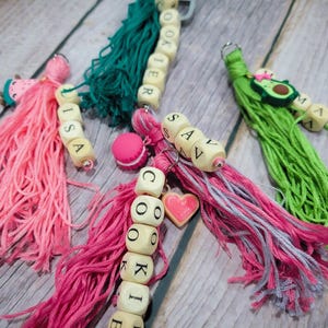 May include: Three decorative keychains featuring colorful yarn tassels in pink, dark teal, and lime green. Each keychain includes wooden letter beads spelling out names or words like LISA, COOKIE, and SAV, along with small charms shaped like food items.