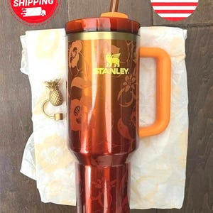 May include: A metallic orange Stanley tumbler featuring a floral pattern, an orange handle, and a matching lid with a straw. Beside the tumbler sits a small gold-colored pineapple-shaped charm. The tumbler displays the Stanley brand logo.