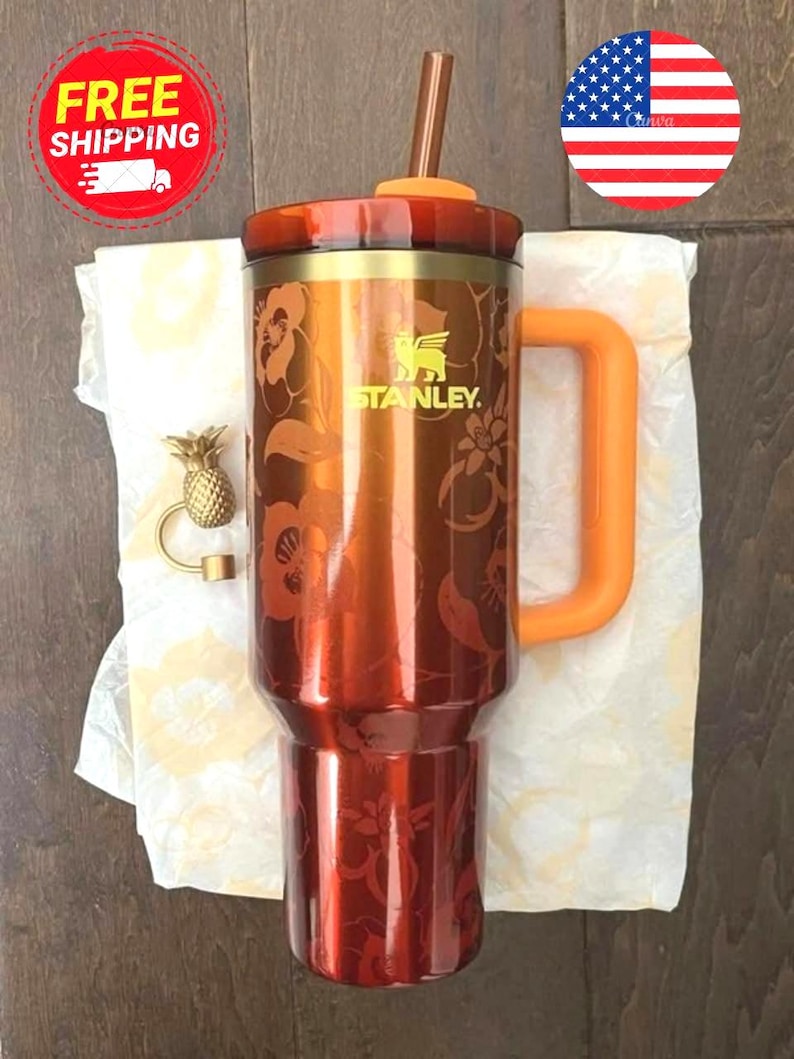 May include: A metallic orange Stanley tumbler featuring a floral pattern, an orange handle, and a matching lid with a straw. Beside the tumbler sits a small gold-colored pineapple-shaped charm. The tumbler displays the Stanley brand logo.
