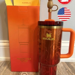 May include: A metallic orange Stanley Quencher H2.0 tumbler with a floral pattern, a gold-tone handle, and a matching orange lid. The tumbler includes a straw topped with a gold-colored pineapple charm and is displayed next to its orange box.