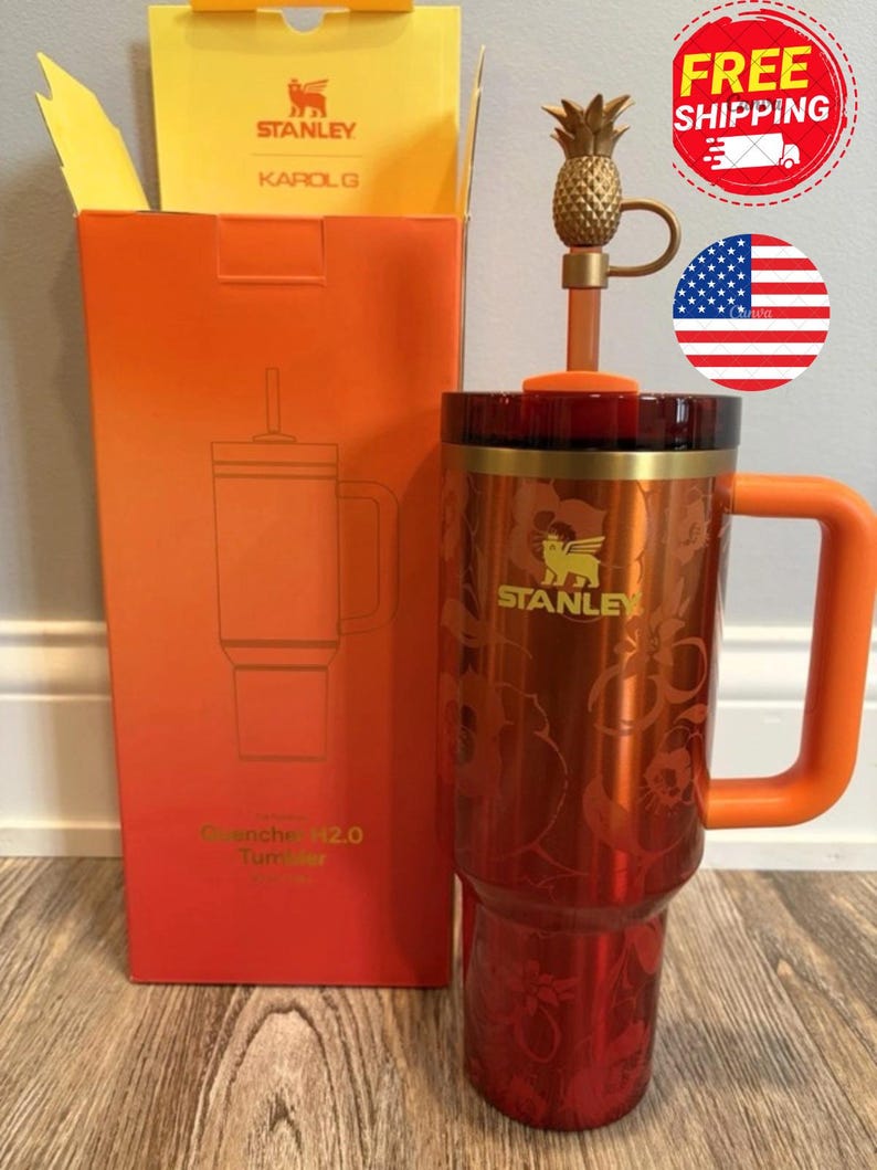 May include: A metallic orange Stanley Quencher H2.0 tumbler with a floral pattern, a gold-tone handle, and a matching orange lid. The tumbler includes a straw topped with a gold-colored pineapple charm and is displayed next to its orange box.