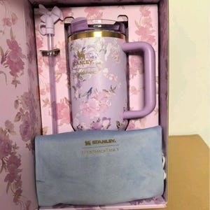 May include: A lavender Stanley tumbler with a floral pattern, a gold rim, and a matching handle. The set includes a straw with a bow topper and a blue velvet pouch, both featuring the Stanley and LoveShackFancy logos, all housed in a floral box.