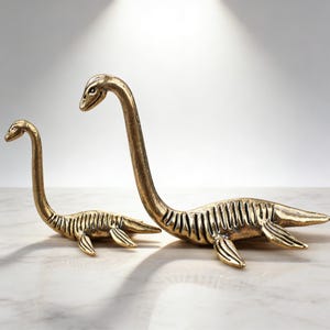 May include: Two brass-colored sculptures of plesiosaurs, one large and one small, are displayed on a white surface. The sculptures have long necks, small heads, and flipper-like appendages. The larger sculpture is approximately 12 inches long.