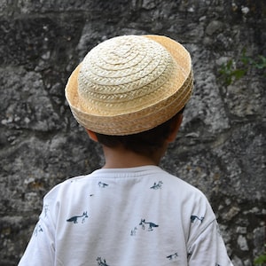 May include: A light beige straw hat with a rounded crown and a short, upturned brim, worn with a white short-sleeved shirt featuring a repeating pattern of small, dark-colored fox illustrations across the fabric.