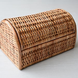 May include: A rectangular woven wicker basket with a rounded, arched lid. The basket features a natural light brown finish and a tight, interlaced weave pattern throughout the body and the curved top, creating a sturdy storage container.