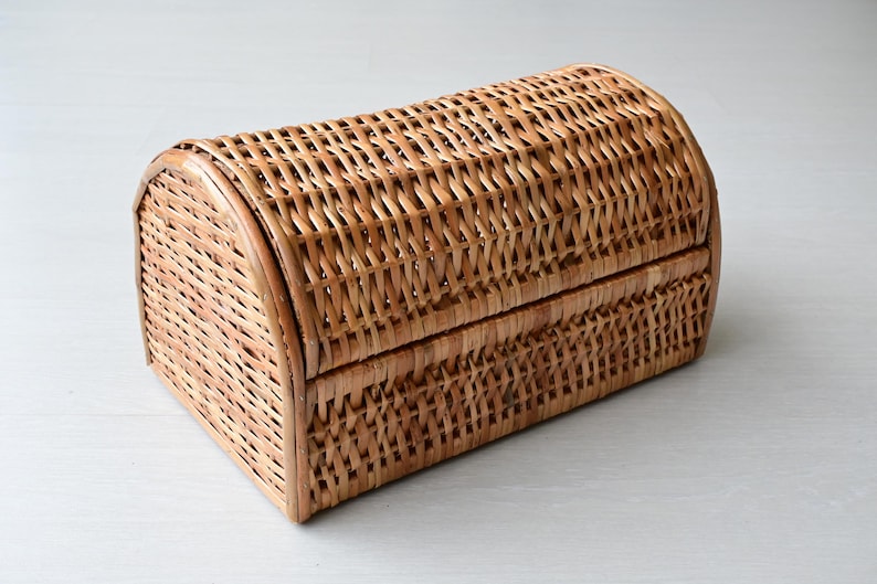 May include: A rectangular woven wicker basket with a rounded, arched lid. The basket features a natural light brown finish and a tight, interlaced weave pattern throughout the body and the curved top, creating a sturdy storage container.