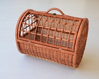 Könnte beinhalten: Ein brauner, geflochtener Weidenkorb als Tiertransportbox in zylindrischer Form. Die Box verfügt über einen aufklappbaren Deckel mit Metallverschluss, einen geschwungenen Tragegriff oben und ein offenes Gittermuster zur Belüftung.