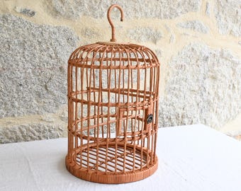 Könnte beinhalten: Ein zylindrischer Vogelkäfig aus hellbraunem, geflochtenem Rattan- oder Weidenmaterial. Die Struktur besteht aus vertikalen Stäben, horizontalen Stützringen, einem kreisförmigen Boden und einem gebogenen Aufhängehaken an der Oberseite.