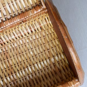 May include: A woven wicker basket featuring a light tan natural finish with a curved structure. The basket is constructed with thin, flexible strips of material interlaced in a tight pattern, framed by a solid, smooth, darker brown wooden edge.