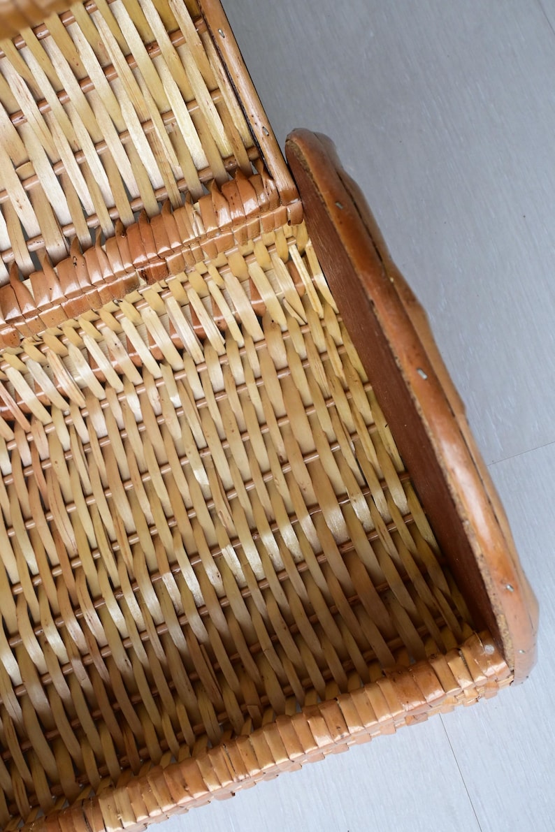 May include: A woven wicker basket featuring a light tan natural finish with a curved structure. The basket is constructed with thin, flexible strips of material interlaced in a tight pattern, framed by a solid, smooth, darker brown wooden edge.