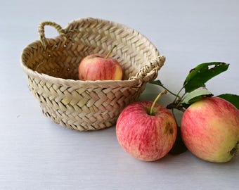 Könnte beinhalten: Ein runder, geflochtener Strohkorb mit zwei kleinen Henkeln, der einen rot-gelben Apfel enthält. Zwei weitere rot-gelbe Äpfel mit grünen Blättern an einem Stiel liegen neben dem Korb auf einer hellen Oberfläche.