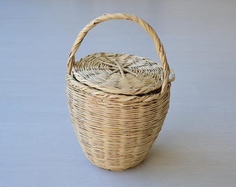 Könnte beinhalten: Ein hellbrauner, geflochtener Weidenkorb mit bauchigem Körper, rundem Deckel und einem einzelnen, bogenförmigen Henkel. Der Korb weist ein enges, horizontales Flechtmuster am Körper und ein strahlenförmiges Muster auf dem Deckel auf.
