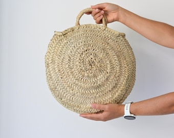 Könnte beinhalten: Eine runde, geflochtene Strohtasche in natürlichem Beigeton mit einem durchgehenden Spiralmuster. Die Tasche verfügt über einen einzelnen, stabilen geflochtenen Griff an der Oberseite, der aus getrockneten Pflanzenfasern besteht.