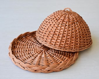 Könnte beinhalten: Eine hellbraune, geflochtene Korbhaube in Kuppelform mit einem kleinen Griff an der Oberseite, kombiniert mit einem passenden runden, geflochtenen Korbtablett. Das Set weist ein durchgehendes Gittermuster aus natürlichem Material auf.