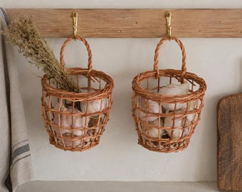 May include: Two light brown woven wicker baskets with arched handles hanging from brass hooks on a wooden rail. The baskets contain white garlic bulbs, and one basket also holds a bundle of dried herbs. The baskets feature an open grid design.