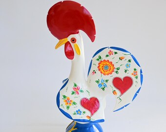 Könnte beinhalten: Eine weiße Hahn-Figur aus Keramik mit einem markanten roten Kamm und Kehllappen, einem gelben Schnabel und blau bemalten Rändern. Körper und Schwanz sind mit handgemalten Blumenmustern in Orange, Gelb und Grün sowie zwei roten Herzen verziert.