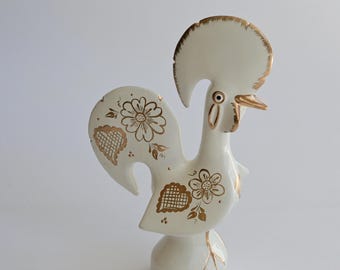 Könnte beinhalten: Eine weiße Hahn-Figur aus Keramik mit goldfarbenen Bemalungen. Das Design umfasst florale Muster und gitterartige Herzformen auf den Schwanzfedern sowie goldene Akzente an Kamm, Schnabel und Augenpartie des Vogels.