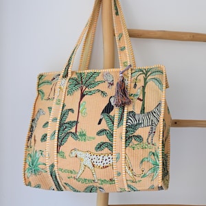 May include: A tote bag made of peach-colored fabric with a quilted texture, featuring a repeating print of jungle animals including a zebra, cheetah, and toucan among palm trees. The bag has two matching fabric handles and a decorative tassel.