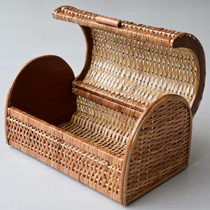 May include: A rectangular woven wicker basket featuring a hinged, arched lid that opens upward. The basket is constructed from light brown natural reeds with a cross-weave pattern on the sides and a solid wooden base and side panels.