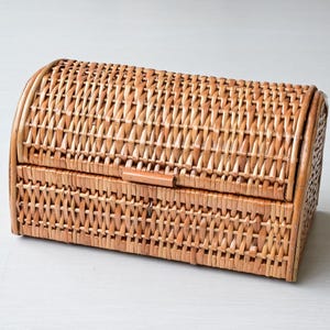 May include: A rectangular woven wicker storage basket with a rounded, hinged lid. The basket features a light brown natural finish and a tight, consistent weave pattern throughout the body and the curved top, providing a structured storage solution.