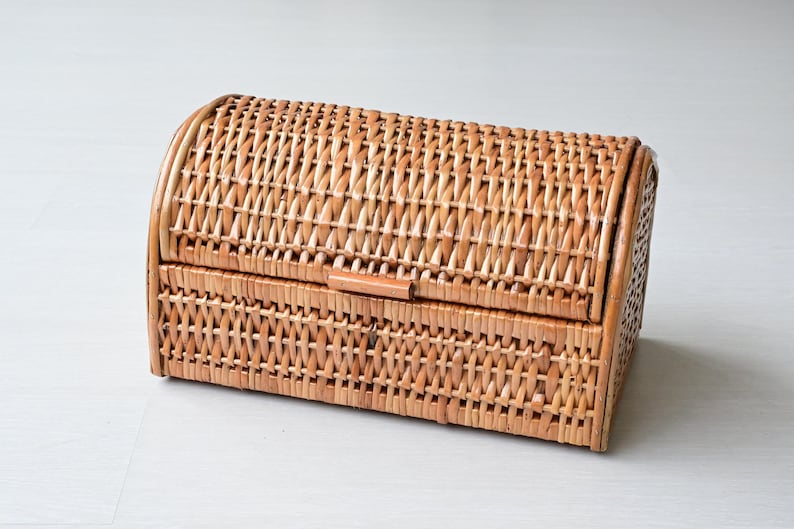 May include: A rectangular woven wicker storage basket with a rounded, hinged lid. The basket features a light brown natural finish and a tight, consistent weave pattern throughout the body and the curved top, providing a structured storage solution.