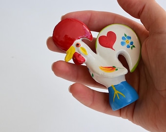 Könnte beinhalten: Eine kleine Hahn-Figur aus Keramik mit weißem Körper, leuchtend rotem Kamm, gelbem Schnabel und blauem Sockel. Der Schwanzbereich ist mit einem gemalten roten Herz und einer kleinen blauen Blume verziert, während eine gelbe Chilischote seitlich abgebildet ist.
