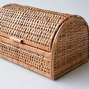 May include: A rectangular wicker basket with a rounded, arched lid. The basket is constructed from light brown woven reeds in a cross-hatch pattern. It features a hinged top cover that extends over the front, secured by a small horizontal latch.