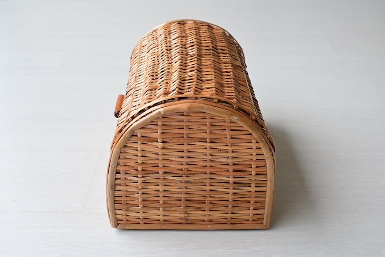 May include: A light brown woven wicker bread box with a semi-cylindrical arched top and a rectangular base. The basket features a horizontal weave pattern throughout, with a small wooden handle attached to the side for opening the curved lid.