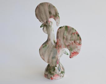 Könnte beinhalten: Eine Hahn-Figur aus Keramik mit einer glänzenden weißen Grundglasur und abstrakten, vermischten Pinselstrichen in gedecktem Grün, zartem Rosa und Grau. Die Skulptur steht auf einem runden Sockel und hat einen hohen, aufrechten Kamm.
