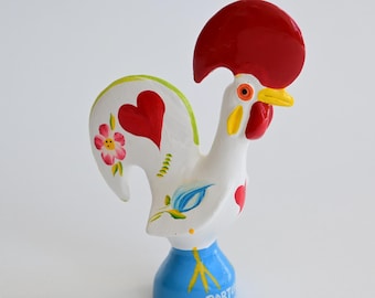 Könnte beinhalten: Eine weiße Hahn-Figur aus Keramik mit einem großen roten Kamm, einem gelben Schnabel und einem roten Kehllappen. Der Körper ist mit handgemalten roten Herzen, einer rosa Blume und blauen Blattmotiven verziert. Der blaue Sockel trägt den Text PORTUGAL.