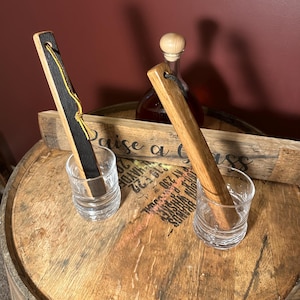 May include: Two wooden muddlers, one with a black top and yellow string, rest in clear shot glasses. A bottle with a wooden stopper and a wooden sign that says "Raise a Glass" are in the background. The items are on a wooden barrel.