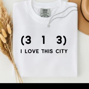 Puede incluir: Camiseta blanca Comfort Colors con el texto negro "(3 1 3) I LOVE THIS CITY". También se ve un collar plateado con un colgante de estrella y un sombrero beige.