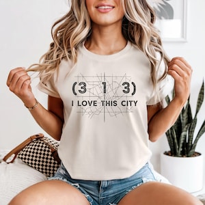 Pode incluir: Camiseta creme com o texto preto "(3 1 3) I LOVE THIS CITY". O design inclui um contorno de mapa estilizado. A camiseta é uma peça de roupa casual.