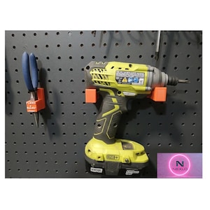 Può includere: Un avvitatore a impulsi Ryobi giallo e nero e delle pinze blu sono montati su una pannello forato grigio scuro utilizzando supporti arancioni. L'avvitatore ha un'impugnatura nera e una batteria One+. Le pinze sono in un supporto arancione.