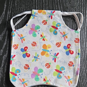 May include: A small, white apron with an elastic strap, patterned with colorful butterflies, dragonflies, and ladybugs. The insects are in shades of red, yellow, blue, green, and purple. Dotted lines connect the insects. The apron is likely for a child.