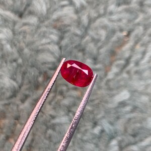 Könnte beinhalten: Ein oval geschliffener Rubin von tiefroter Farbe, gehalten von einer silbernen Pinzette. Die Facetten des Edelsteins sind sichtbar. Die Pinzette befindet sich vor einem weichen, strukturierten, grauen Hintergrund, der die Farbe und Klarheit des Rubins hervorhebt.