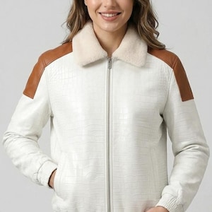Puede incluir: Una chaqueta tipo bomber blanca con un patrón texturizado de relieve de cocodrilo, un cuello de piel sintética color crema y paneles de cuero marrón claro en los hombros. La chaqueta incluye una cremallera plateada, puños y dobladillo acanalados.