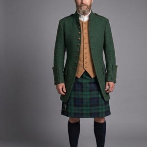 Puede incluir: Chaqueta de lana verde con botones dorados, usada sobre un chaleco beige y una camisa blanca con cuello. El atuendo incluye un kilt tartán tradicional en verde y azul, calcetines altos negros y zapatos de cuero marrón. El fondo es gris liso.