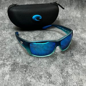 May include: Blue and teal sunglasses with mirrored lenses and a black case. The sunglasses have a dark blue frame with teal accents and a blue logo on the case. The lenses reflect a vibrant blue color.