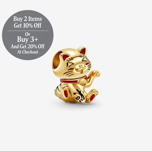 Puede incluir: Un charm de Maneki Neko dorado con detalles en rojo. La figura del gato de la suerte está sentada con una pata levantada. El charm tiene detalles negros y está sobre un fondo blanco.