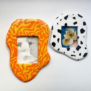 Op de afbeelding: Twee unieke, handgemaakte fotolijsten. Eén lijst is oranje en geel met een vlamachtig patroon, met een potloodschets. De andere is wit met zwarte vlekken, met een foto van bloemen. Beide lijsten hebben onregelmatige, organische vormen.