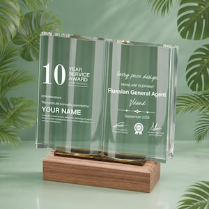 May include: A clear glass award in the form of an open book, set on a wooden base. The left side displays "10 YEAR SERVICE AWARD" and "YOUR NAME". The right side reads "Imrg pace design" and "Russian General Agent".