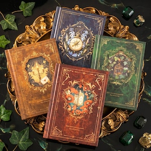 Puede incluir: Colección de cinco diarios decorativos con cubiertas ornamentadas. Los diarios presentan diseños que incluyen una brújula, una jaula para pájaros y motivos florales. Las cubiertas son en tonos marrón, verde y azul, con detalles dorados. Los diarios están colocados en una bandeja dorada.