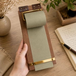 May include: A wooden notepad with a roll of green paper. The notepad has brass hardware and brown leather accents. The notepad is held by a person's hand.